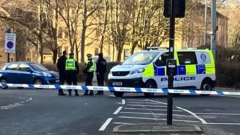BBC Police officers standing next to a police van on a street. There is blue and white police tape in the foreground, with another car behind the police van on an empty road.
