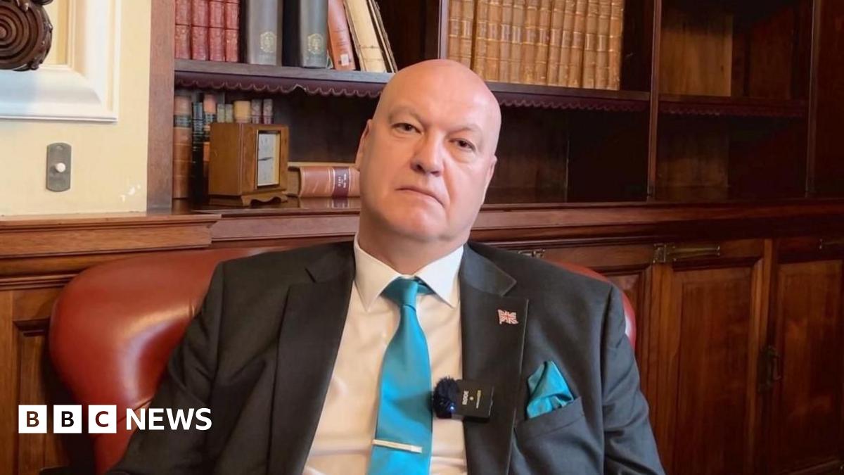 A brown NC500 sign post which says it is 27 miles to Ullapool next to a country road with hills in the background.