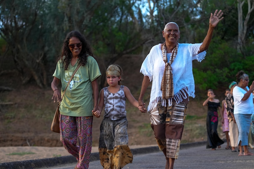Two adults and a child hold hands, smiling and waving as they walk.