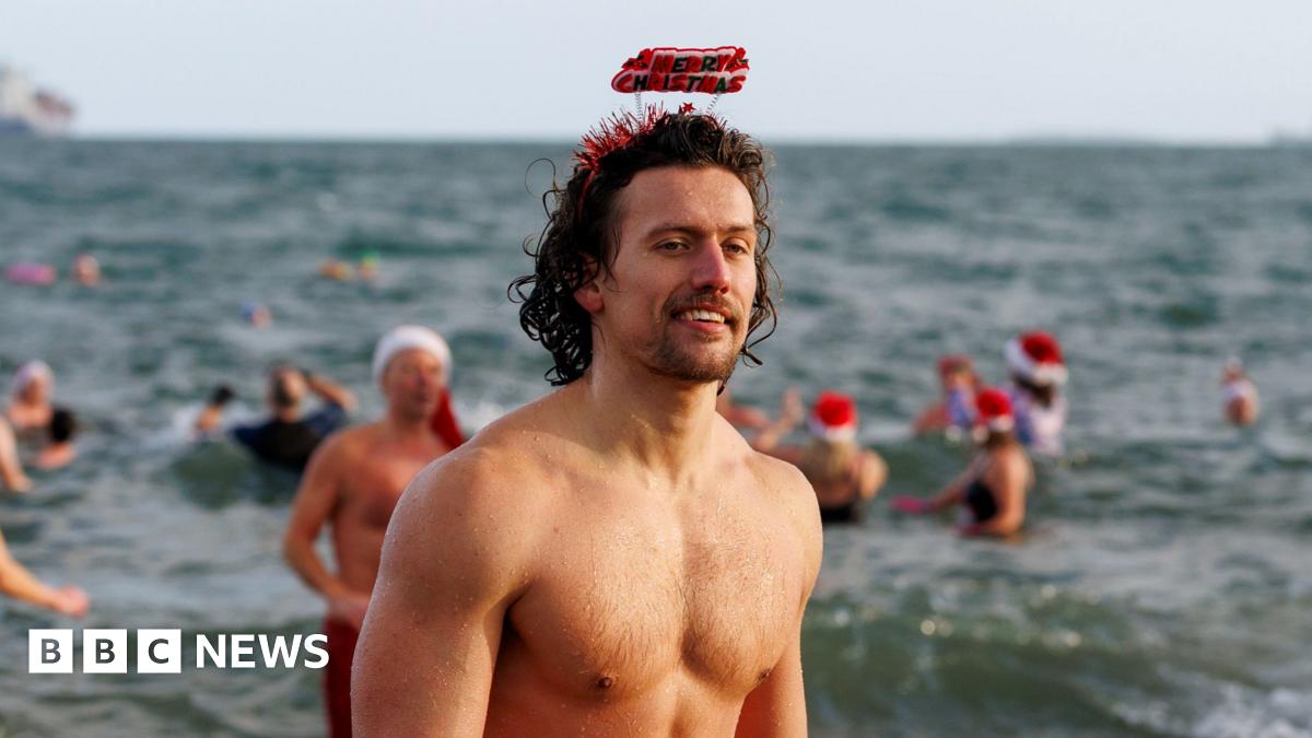 Eight woman stand and smile at a camera. They are all wearing red Santa hats, that have white headbands. The woman on the far right wears a blow up peach-coloured Turkey costume.