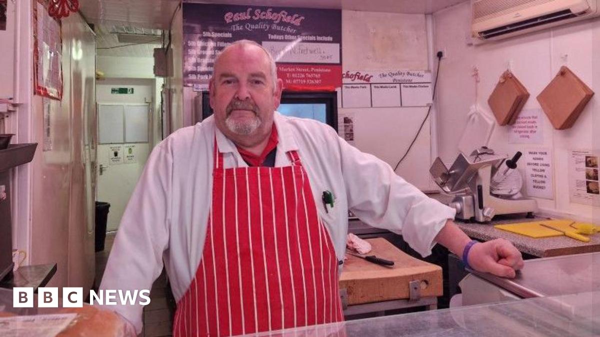 Butcher Paul Schofield stands behind his counter in The Quality Butcher on Market Street in Penistone. He is wearing a red and white striped butcher's apron. A yellow chopping board and knife are to the right of his hand, which is resting on the glass counter.