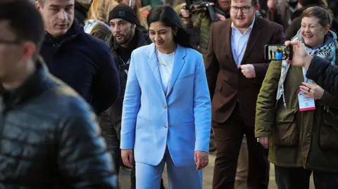 Getty Images Zarah Sultana in a light blue suit walks through a crowded area, surrounded by people holding cameras, microphones, and smartphones. Several individuals have press badges and lanyards, and the background shows a busy scene with media staff and supporters.