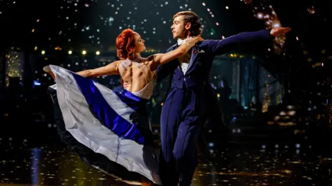 George and Alexis on the dancefloor, he is wearing a blue tailcoat and Alexis is in a shiny dress with a blue skirt. A disco ball is reflecting spots of light all around them.