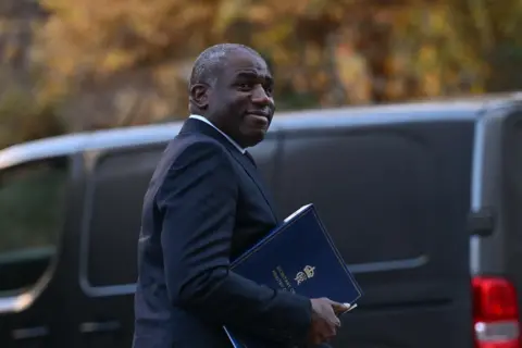 Getty Images David Lammy standing in front of a black van and smiling. He is holding a royal blue folder. 