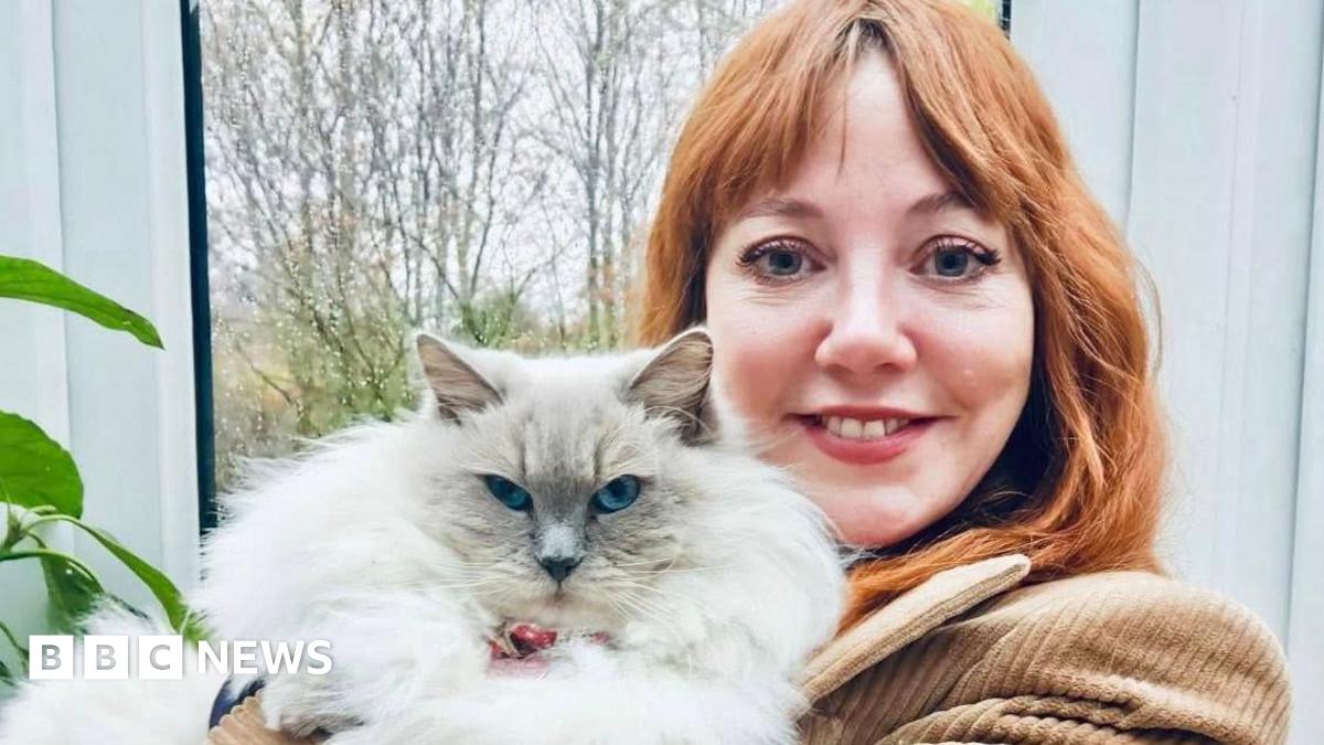 Diane Morgan, pictured holding a large, fluffy white cat which looks directly at the camera. Diane has long red hair and wears a brown corduroy jacket; she is smiling at the camera, standing in front of a window with trees in the background.