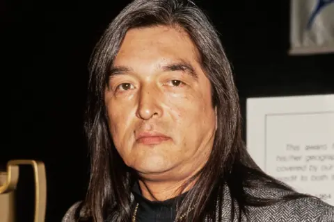 Getty Images Graham Greene has long straight hair and is wearing a patterned blazer over a dark top.