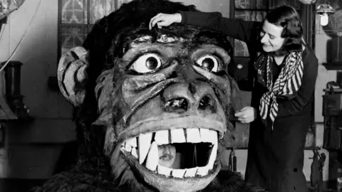 Getty Images A woman stands working on a large monkey head prop for a theatre show. A man is visible standing between the mouth of the prop. 