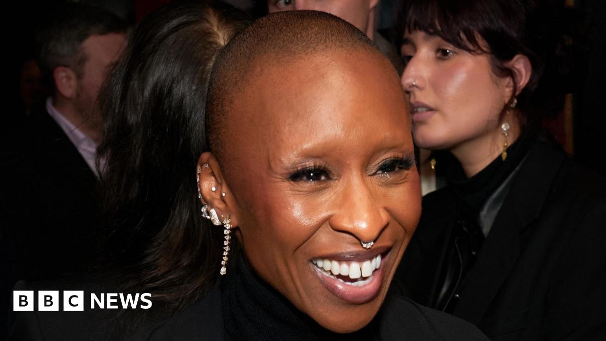 Cynthia Erivo smiling at a Wicked: For Good event, she is wearing several diamond earrings and a diamond nose ring. Her head is shaved.