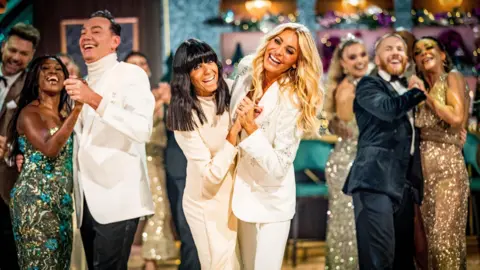 Claudia Winkleman and Tess Daly, both wearing white, join hands as they dance towards the camera, with judges and professional dancers behind them 