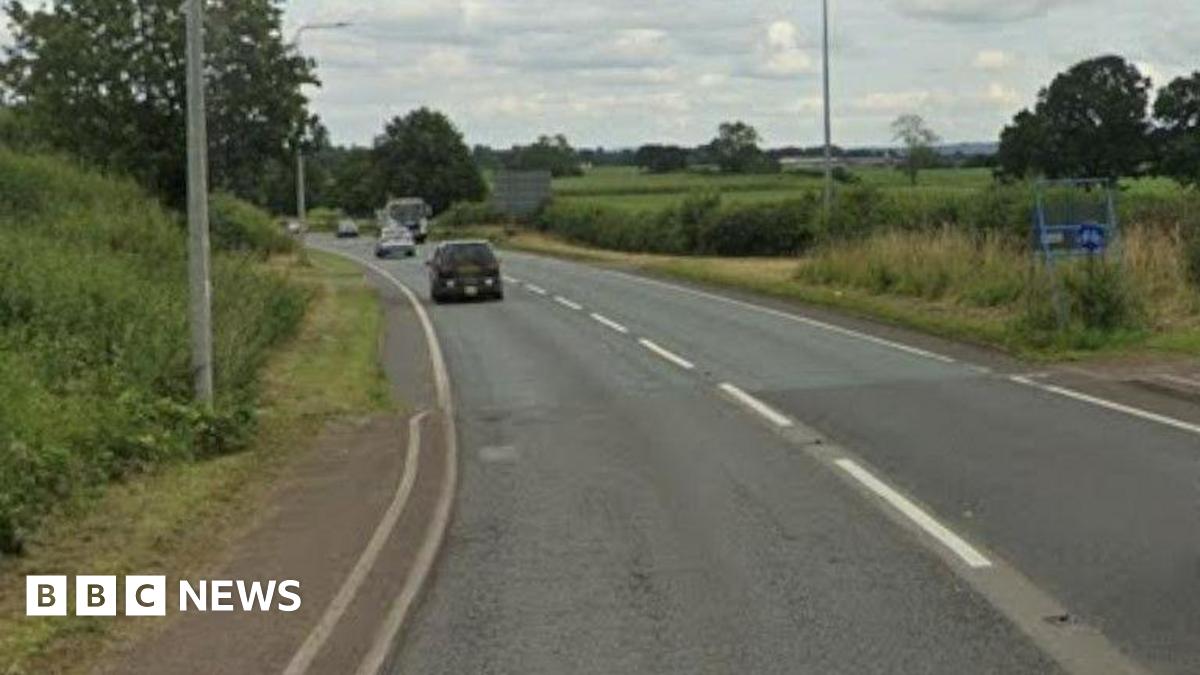 A road, with numerous cars travelling in both directions, is seen curving to the left. There are hedges and fields in the distance and a footpath in the foreground.