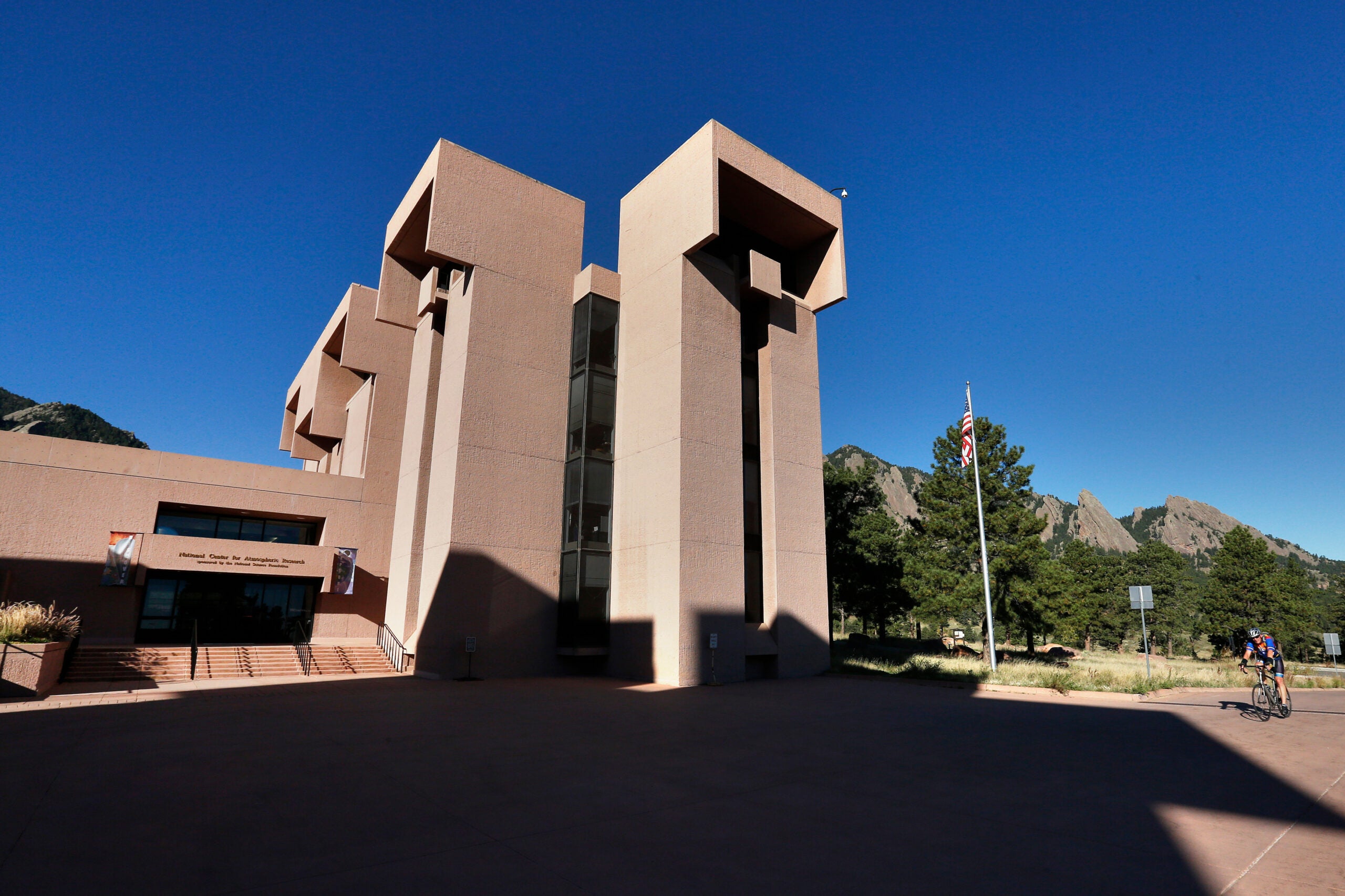 A modern, angular building with tall columns stands under a clear blue sky, with mountains and trees in the background and a cyclist passing by the entrance.