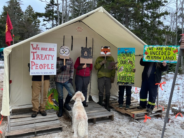 Outside a tent, people with animal masks are standing with "We are all treaty people" banners.