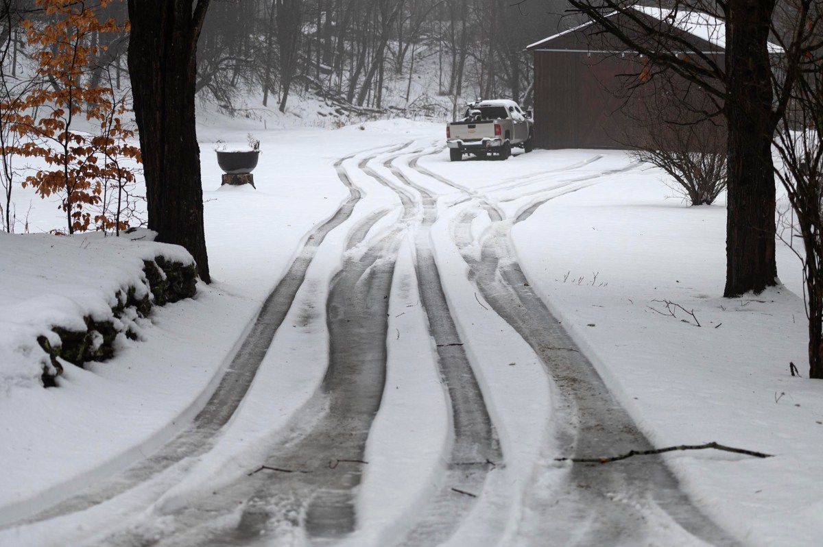 Snow-covered driveway with tire tracks leading to a white pickup truck parked near a barn, surrounded by trees in a winter landscape.