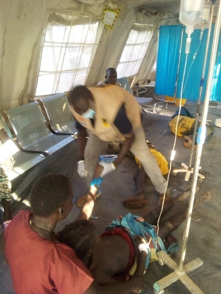 In a blurry photograph, a person in a tan vest and surgical mask bends over to place an IV into the arm of a person who is lying on the floor, mostly undressed. Another person holds them.
