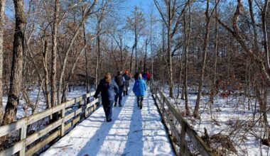 A group of people walking on a snowy wooden boardwalk at Buckhorn State Park through a forest under a clear blue sky.