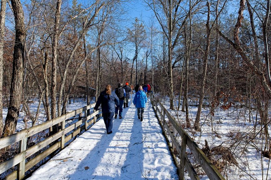 A group of people walking on a snowy wooden boardwalk at Buckhorn State Park through a forest under a clear blue sky.