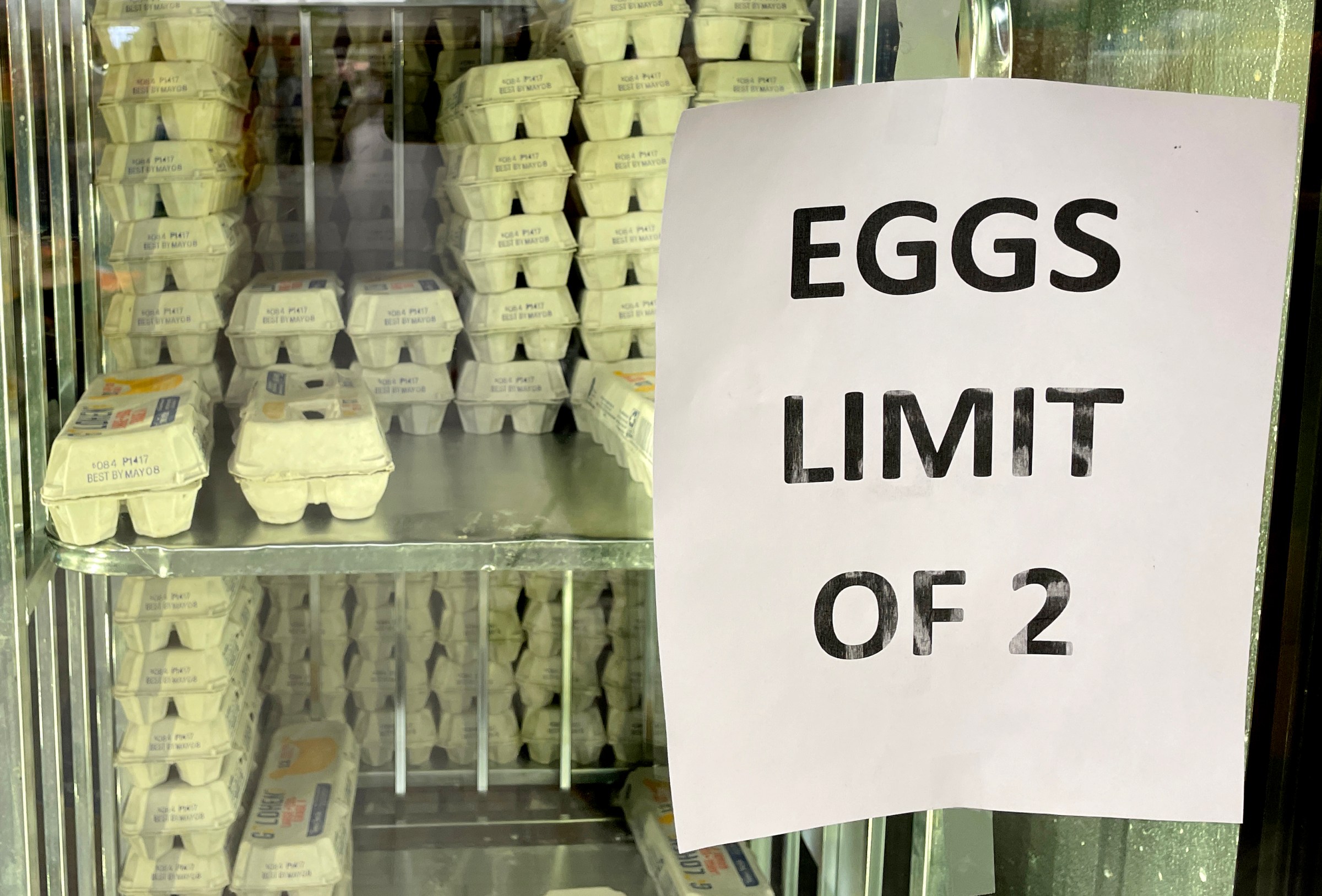 A grocery store cooler full of eggs, with a paper sign reading “Eggs Limit of 2.”