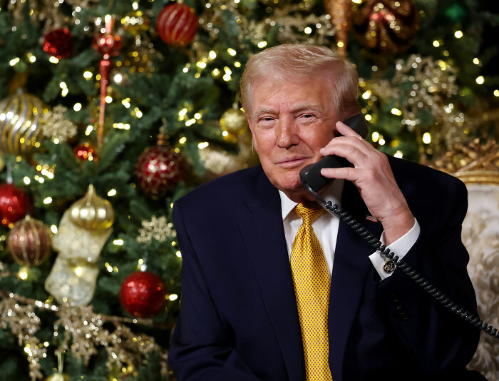 Donald Trump in a dark suit on the phone with a decorated Christmas tree in the background during holiday season.