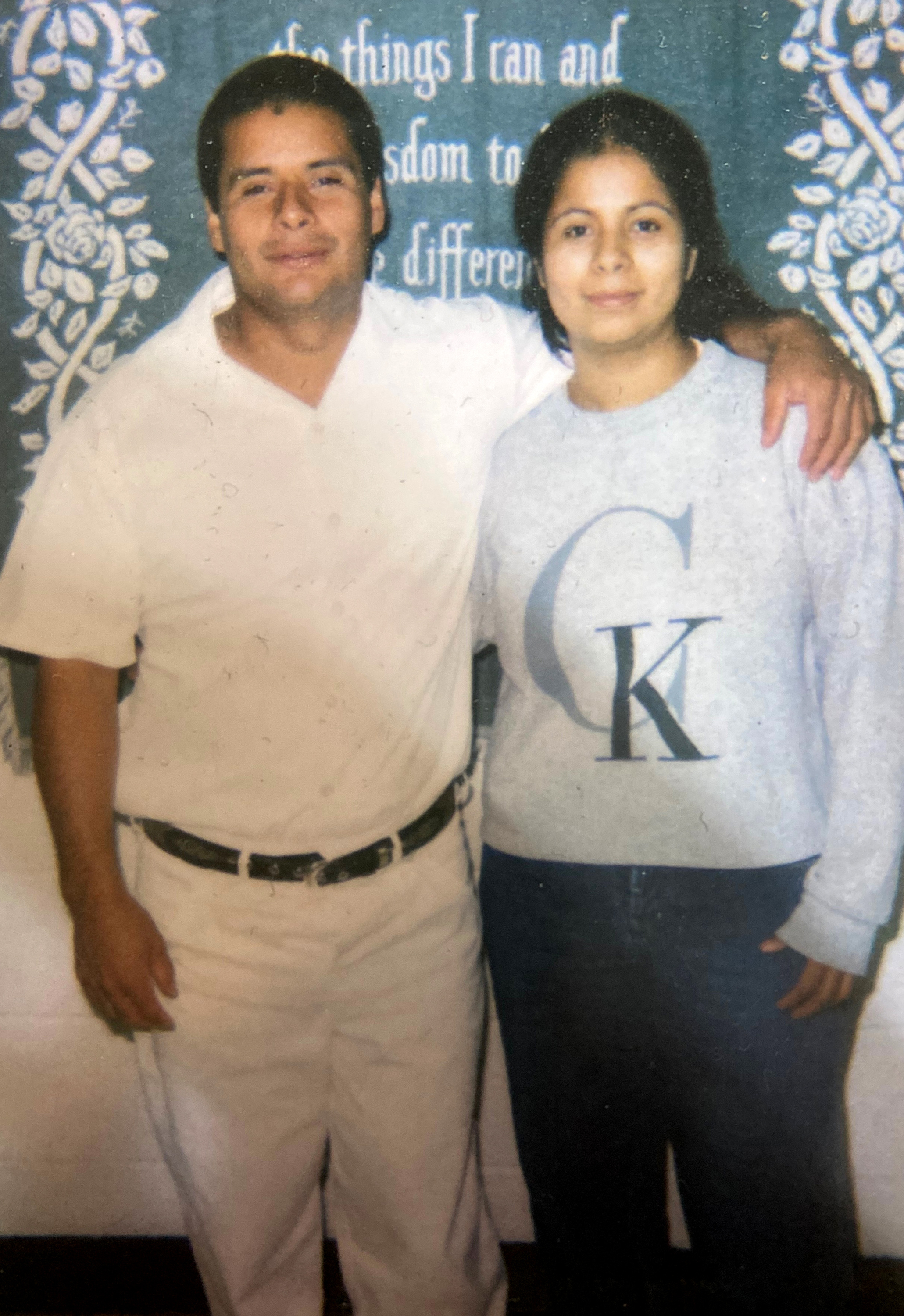 A smiling man wearing white pants and short-sleeve white button-down shirt stands with his arm around a smiling woman wearing a sweatshirt with the Calvin Klein logo. They are standing in front of a tapestry with the words “things I can … wisdom to … difference.”