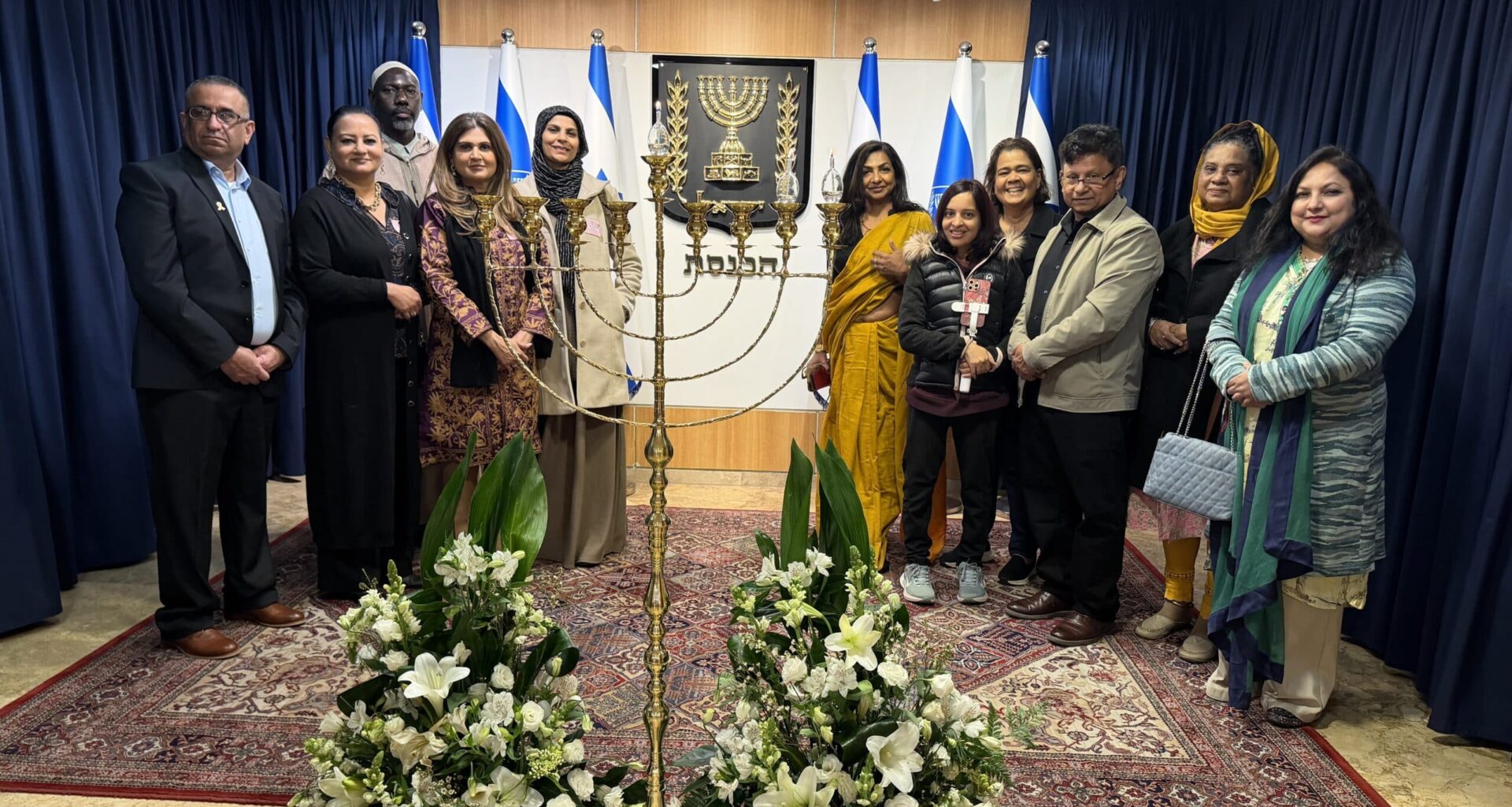 Members of the AMMWEC–CAM delegation stand beside a menorah inside the Israeli Knesset, with Israeli flags and the state emblem in the background.