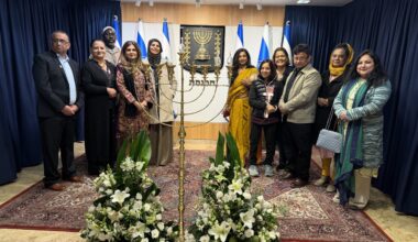 Members of the AMMWEC–CAM delegation stand beside a menorah inside the Israeli Knesset, with Israeli flags and the state emblem in the background.