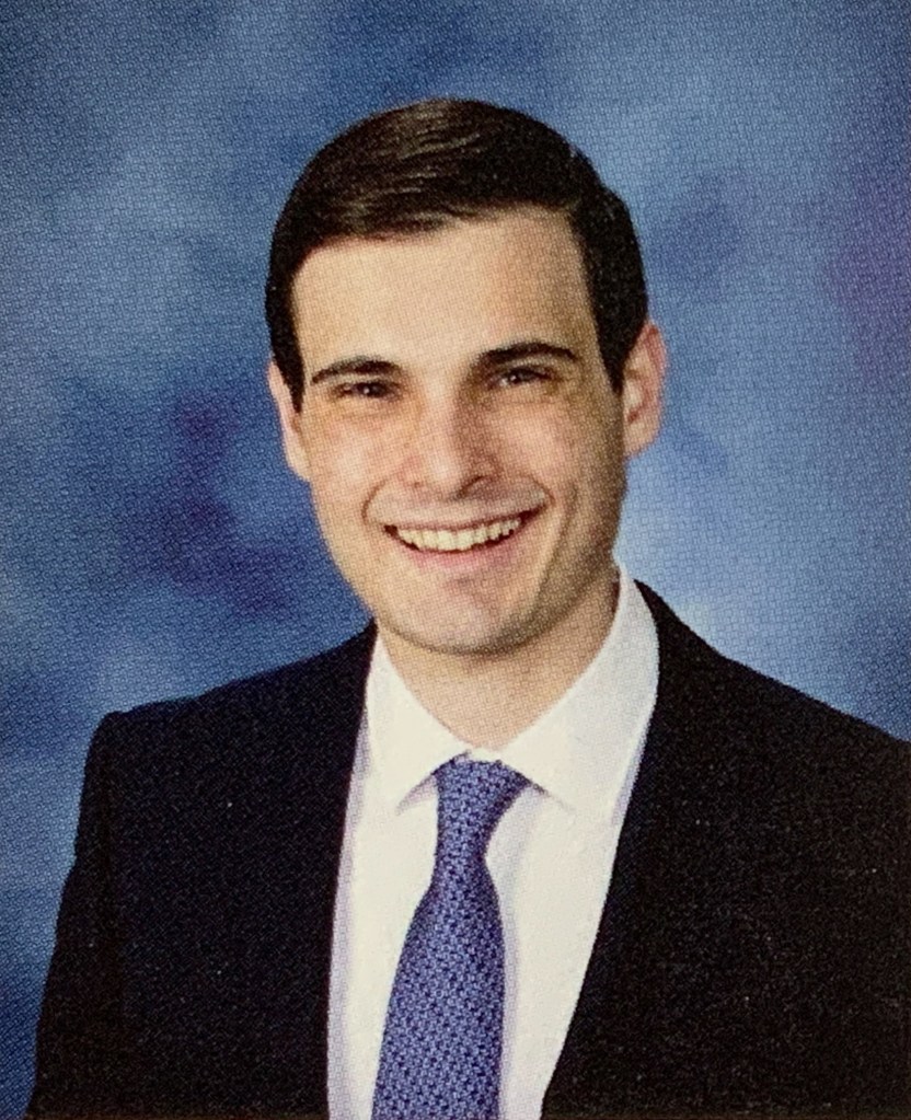 A yearbook photo of a man in a suit looking at the camera, in front of a blue background.
