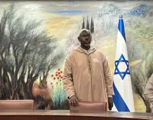 Imam Musa Drammeh stands inside a meeting room in Israel, with an Israeli flag visible behind him.