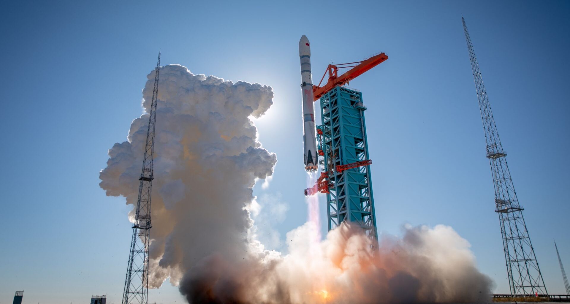 A tall white rocket held up by red scaffolding begins to fire its engines, creating a cloud of white smoke.