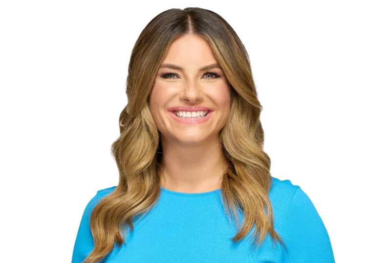 Smiling woman with long, wavy blonde hair wearing a vibrant blue top, against a plain white background.