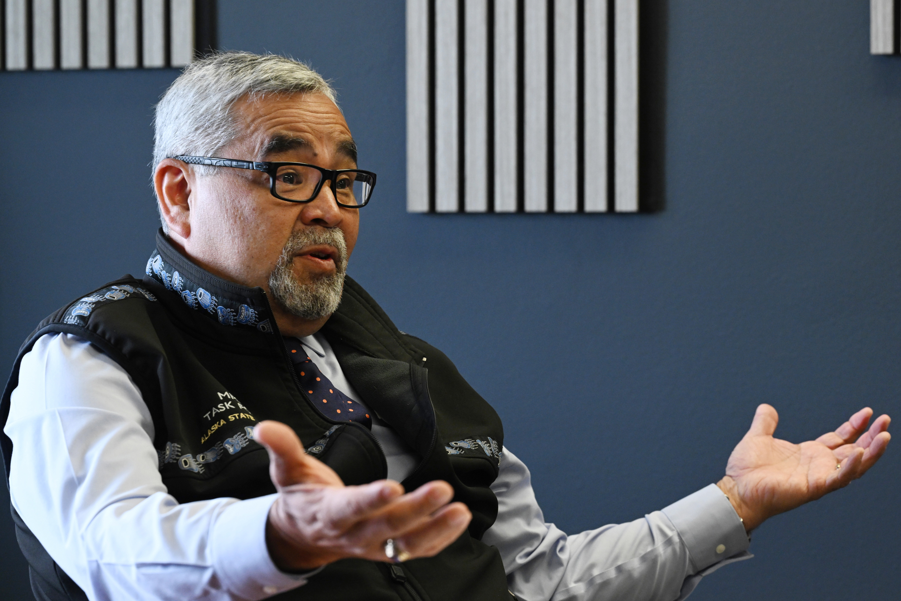 A man with gray hair and a gray moustache and goatee, wearing a black vest with embroidered accents and black framed glasses, raises his hands palm up.