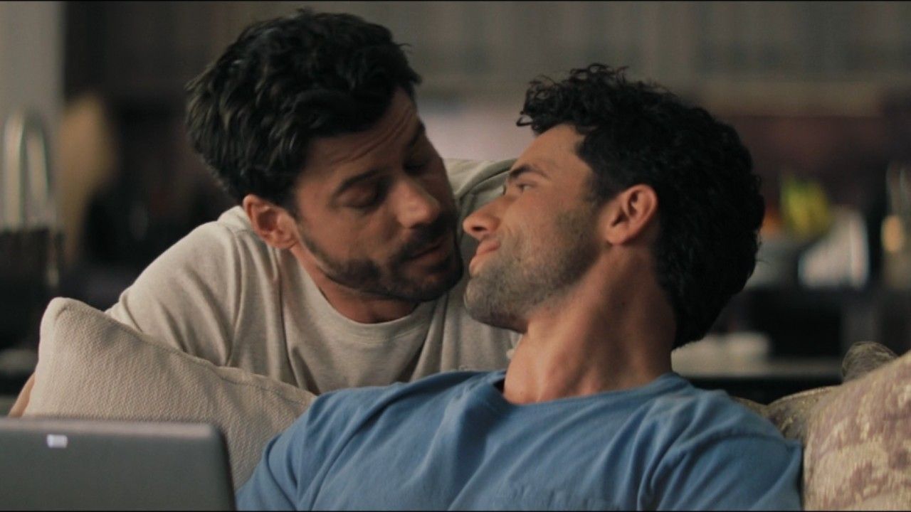 From left to right: Scott leaning against the back of the couch and looking at Kip who is sitting on the couch and looking up at Scott.