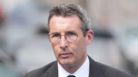 PA Media  Muir is outside. He's wearing a black suit with a white short and a black tie. 