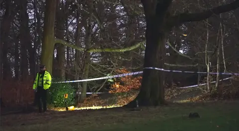 BBC A police officer in a high viz jacket and police tape around a wooded area