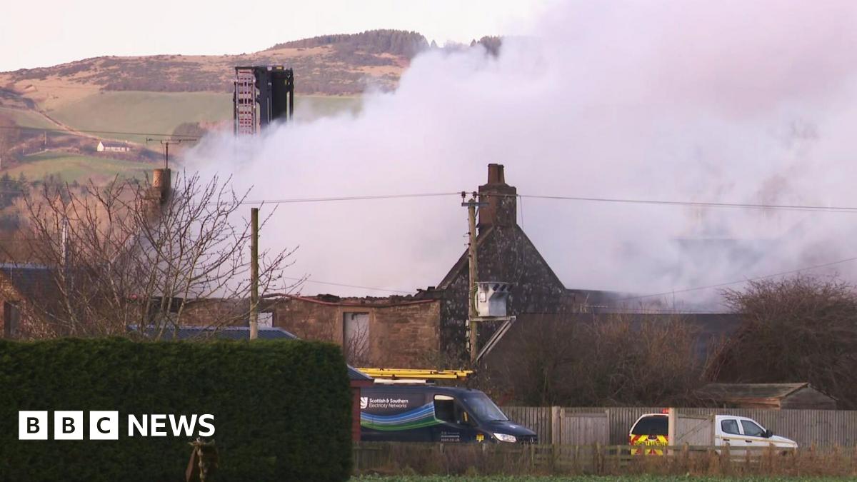 Smoke billows from the remains of a single-storey stone house, which no longer has a roof. In the foreground there is a hedge and a couple of vans, and there are hills and a tower in the background.