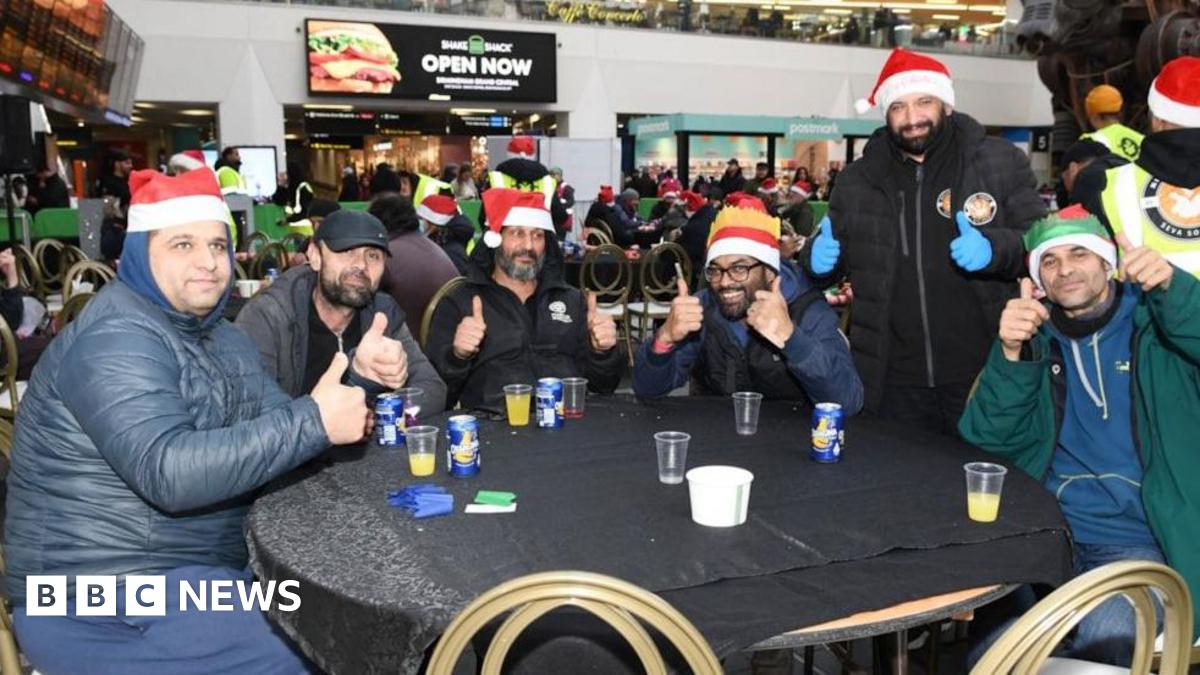 A group of homeless people sat around a table, inside a trains station posing with their thumbs up and smiling at the camera