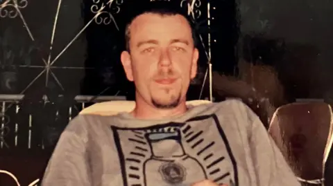 Alison Mark Jenner, pictured by Alison, on one of their holidays. He wears a grey t-shirt with an illustration on the front. 