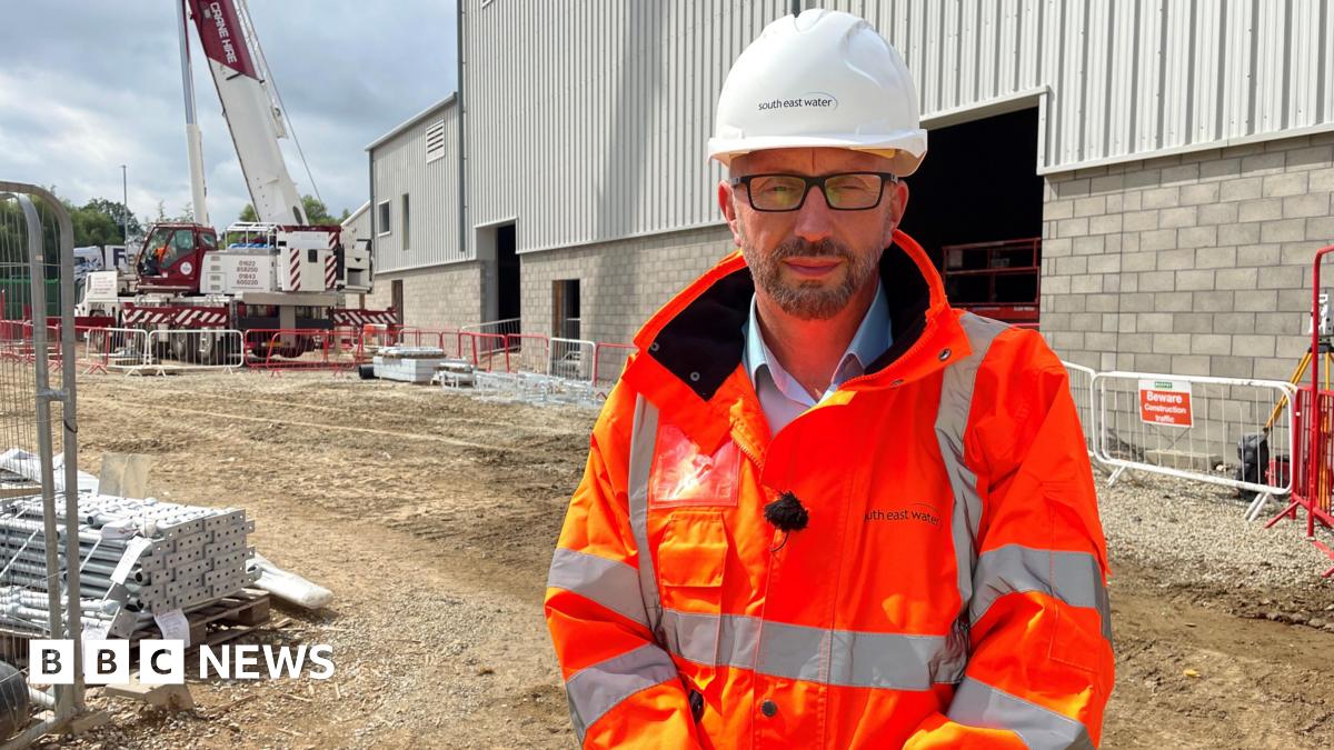 A man wearing an orange hi-vis jacket and a white work and safety hat. He is standing in a construction area.