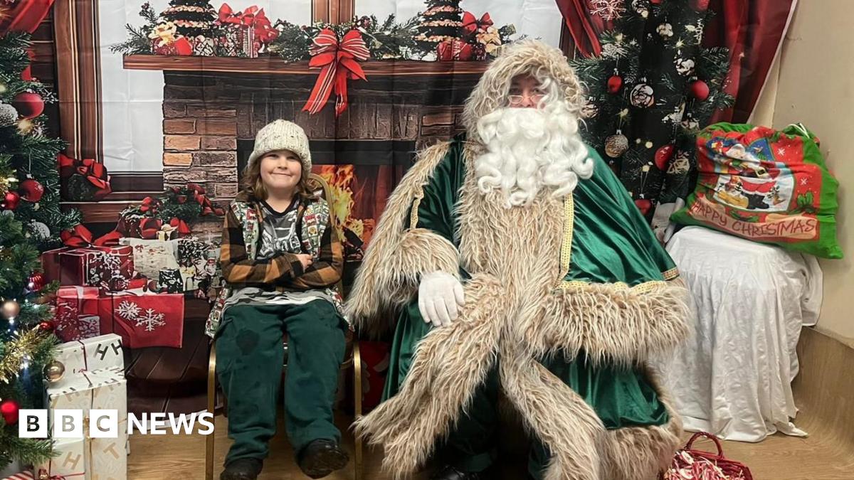 A child wearing a knitted hat and jumper sits beside a man dressed as St Nicholas, who is dressed in a green robe with a long white beard. They are indoors in front of a festive backdrop.