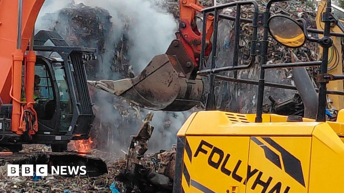 An excavator is being used to dig through a large pile of waste. Orange flames are seen on the ground in the background.