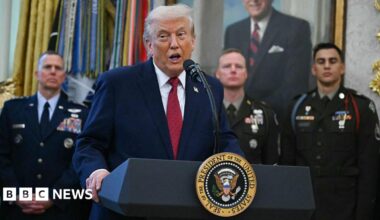 US President Donald Trump wearing a blue suit and red tie in the Oval Office in front of US servicemembers on 15 December.