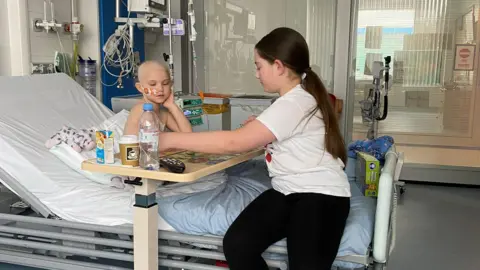 Family Ellie sits on a hospital bed with her little sister Emilia. Emilia is going through chemotherapy and has lost her hair. A table is beside them with a bottle of water and other drinks. They are playing a game together. There is hospital equipment all around them.