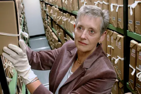 PA Media Dame Stella Rimington is wearing white gloves is handling a large archival box in a narrow aisle lined with shelves full of similar brown archival boxes tied with white strings. The shelves are densely packed, and each box has a visible label with numbers. She appears to be in an organised archive or records storage facility.