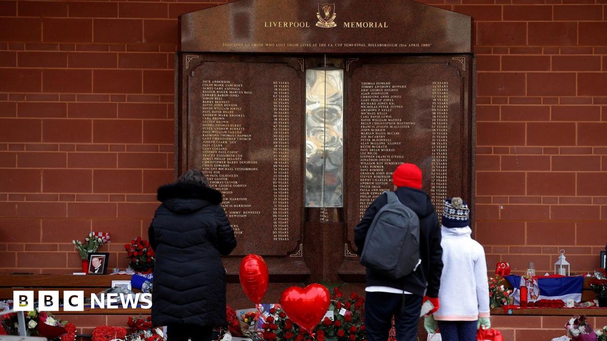 A woman and a man with a child stand in front of the memorial at Anfield that lists the names of those who died. Red heart shaped balloons, flowers, candles and picture frames are beneat the plaque of 97 names.