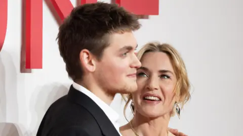 Netflix Joe Anders on the red carpet with his mum Kate Winslet. Winslet is looking up at him proudly and smiling.