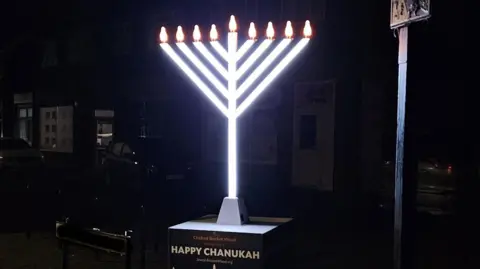 Chabad Lubavitch Bricket Wood A street scene at night with a large menorah , which is the Jewish candelabra, with 9 bulbs on top. The stem and V-shaped prongs are lit up and glowing white. It stands on a black box with the words Chabad Bricket Wood wishes you a Happy Chanukah. Behind it are houses with cars on their driveways. 