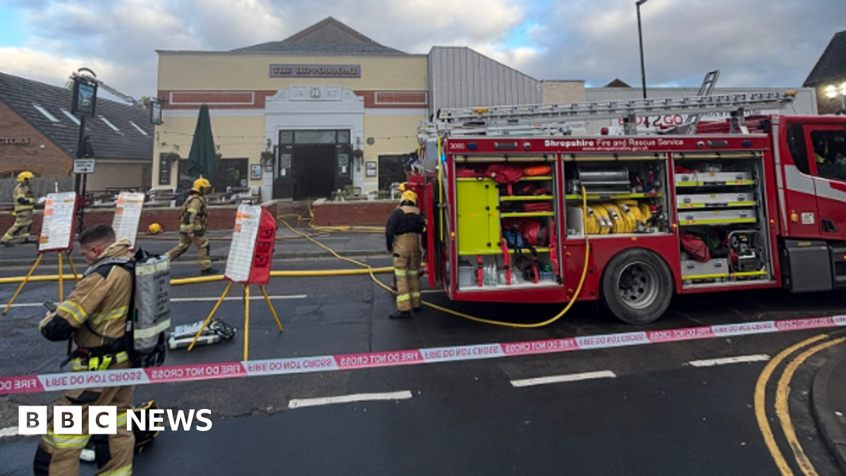 Four firefighters are on the left and in the middle of the photo. A fire engine is to the right and a 'fire do not cross' cordon is in the foreground. A mainly yellow building is in the background.