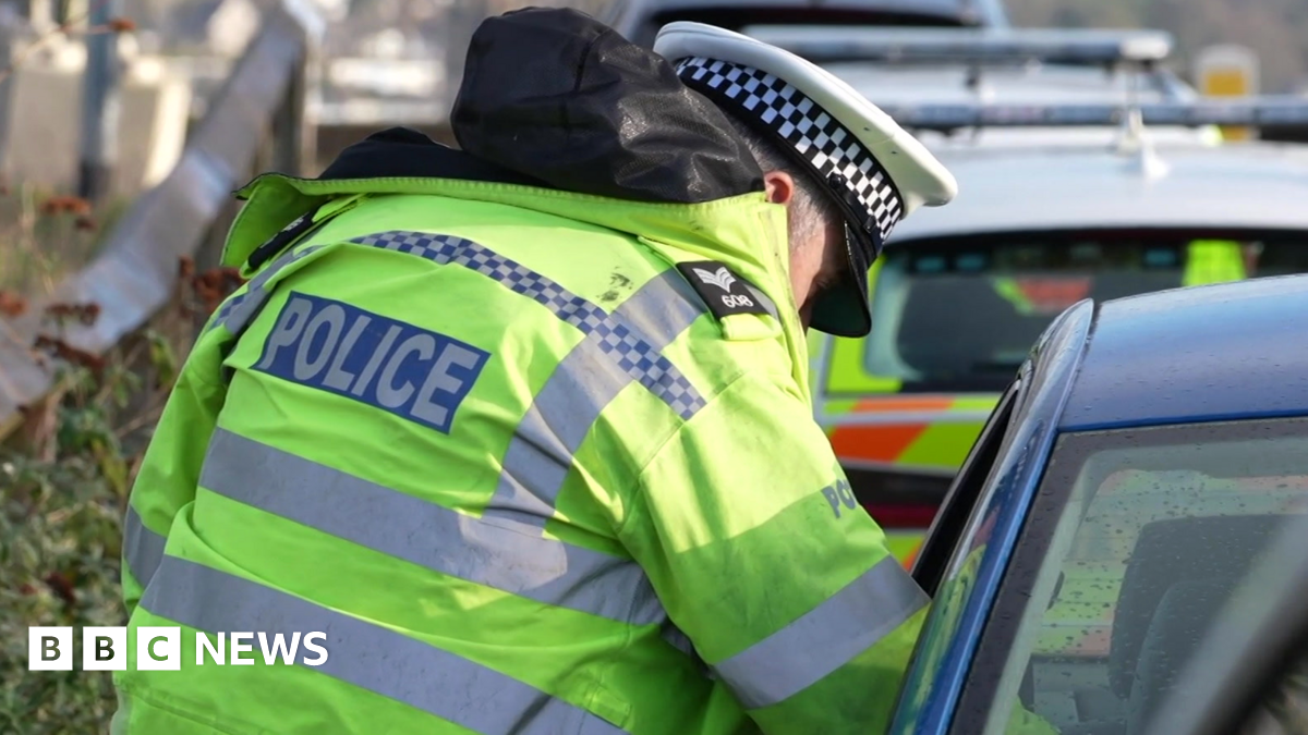 The image shows a roadside scene involving a police officer and a vehicle. The officer is wearing a high-visibility fluorescent yellow jacket with reflective stripes and the word “POLICE” clearly visible on the back. The officer is also wearing a black hood and a white peaked cap with a black-and-white checkered band. The officer is leaning towards the window of a blue car, likely speaking to the driver or checking something inside the vehicle.