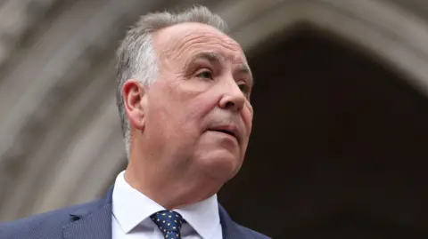 EPA/SHUTTERSTOCK Chris Whitbread looking off to the right of the frame. He is wearing a navy suit jacket, white shirt and blue polka dot tie.
