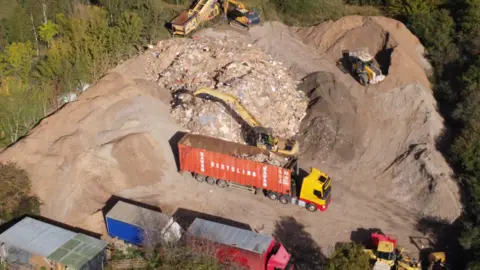 Peopleton Community An aerial view of large piles of earth and mixed debris. Lorries are pictured moving rubble with several other excavator-type vehicles around the area, all surrounded by trees.