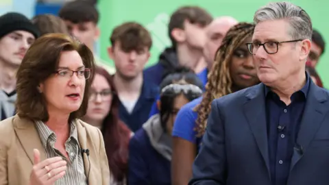 PA Media Eluned Morgan, a woman with brown hair and glasses, speaks in front of a crowd of people while standing next to Sir Keir Starmer, a man with grey hair and glasses.